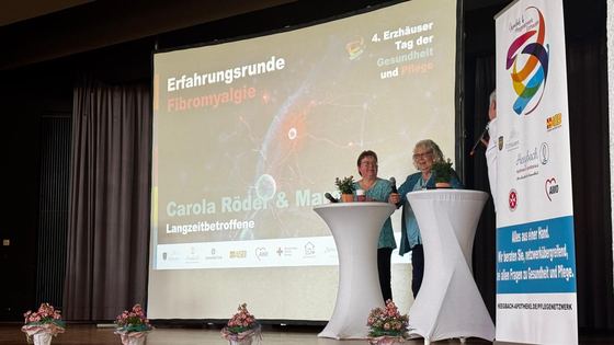 Erfahrungsrunde auf der Bühne. Zwei Personen stehen an Stehtischen, auf der Leinwand ihre Namen. 4. Erzhäuser Gesundheits- & Pflegetag Erzhausen 2026.