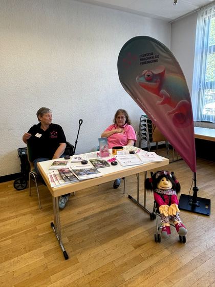 Infostand mit zwei Mitarbeiterinnen, davor ein Maskottchen in einem Kinderrollstuhl. 4. Erzhäuser Gesundheits- & Pflegetag Erzhausen 2026.