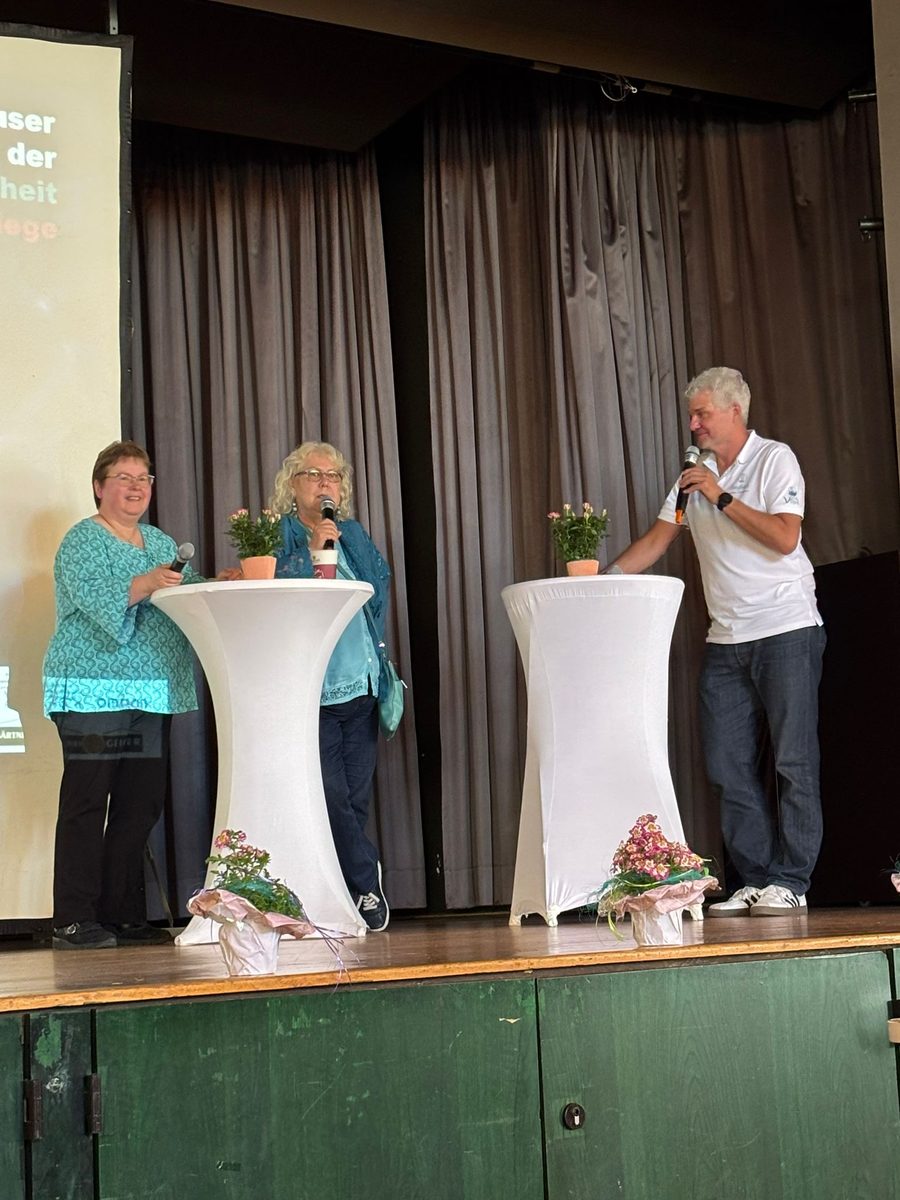 Zwei Frauen und ein Mann stehen mit Mikrofonen an weiß bezogenen Stehtischen auf der Bühne und sprechen miteinander. Auf den Tischen stehen kleine Rosenstöcke. 4. Erzhäuser Gesundheits- & Pflegetag Erzhausen 2026.